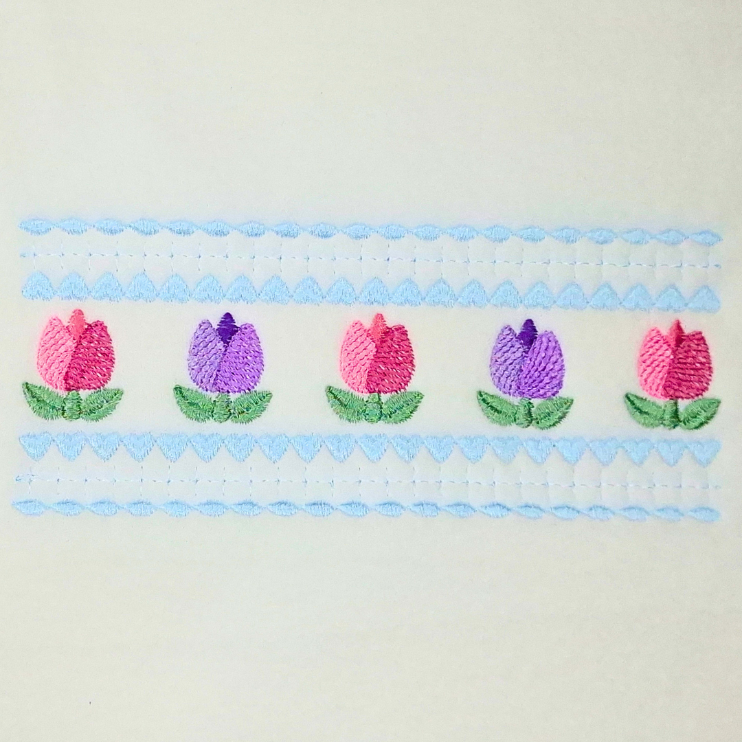 This is an image of a floral inspired machine embroidery design by Stitches & Strokes, featuring a beautiful brightly colored row of tulips in bloom with a delicate lace border. The Tulip Border Machine Embroidery Pattern is perfect for embroidery on Kitchen items like Tea- and Dish Towels, Aprons, and Table Runners.