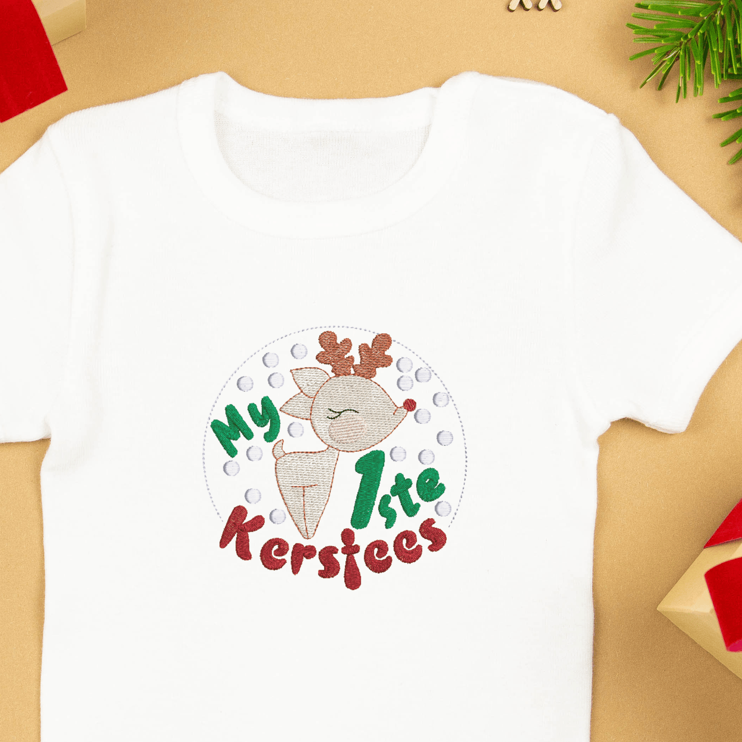 A sweet snow globe Afrikaans quote machine embroidery design for baby's first Christmas with a little baby deer with a red nose and snow flakes. An example of the design on a baby t-shirt. 