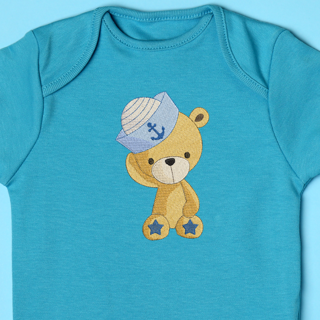 This is an image of a baby bear with a sailor hat machine embroidery design. The baby bear's hat features a little ship's anchor and the baby bear has blue stars on his feet.