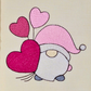 This is an image of a machine embroidery design featuring a little gnome with a pink hat and pink heart shaped balloons. This Gnome machine embroidery design can be used for embroidery on bags, t-shirts, accessories, and gifts for Valentine's Day.