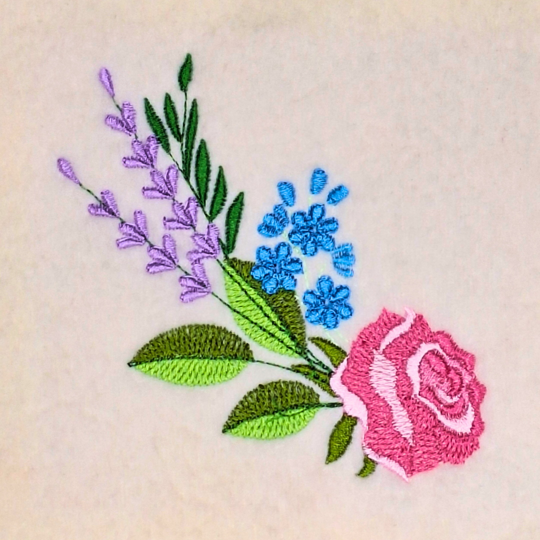 This is an image of a machine embroidery design by Stitches & Strokes, featuring a pink rose with green foliage, purple lavender flowers and blue flowers. This Rose Flower Bouquet Machine Embroidery Design faces in one direction, making it perfect for embroidery on one panel of a jacket or hoodie with a zipper. This design can also be mirrored so that the the design looks the same both panels of the garment.